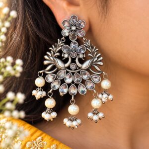 Pair of silver-tone peacock chandelier earrings with clear accents and cream pearl-like drops on a dark background.