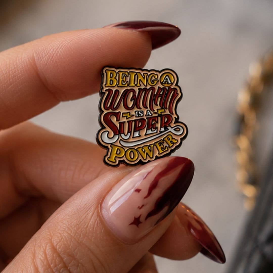 Being a Woman is a Superpower feminist enamel pin held between fingers with dark red manicure