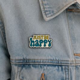 Just Be Happy soft enamel pin in teal and yellow attached to a light blue denim jacket