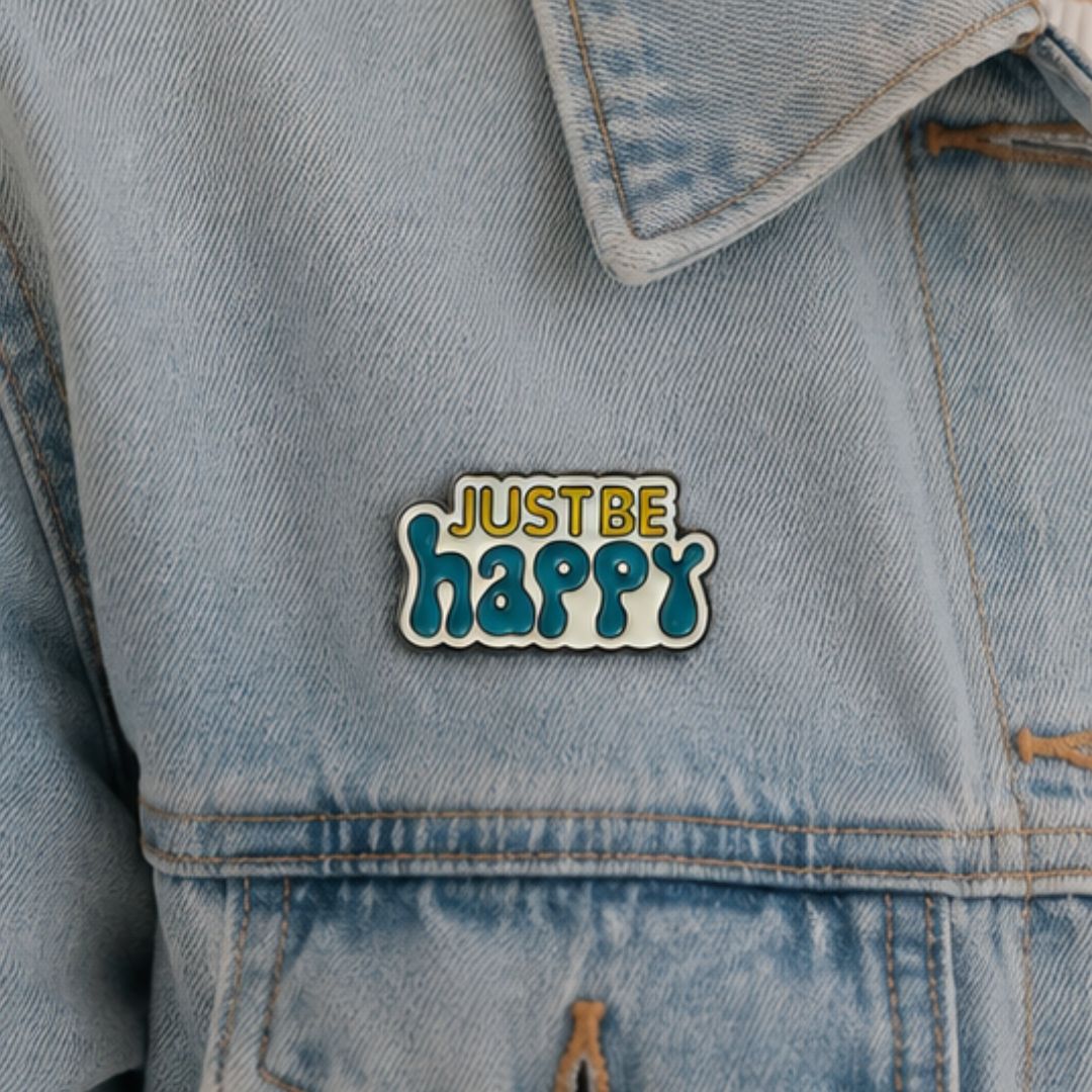 Just Be Happy soft enamel pin in teal and yellow attached to a light blue denim jacket