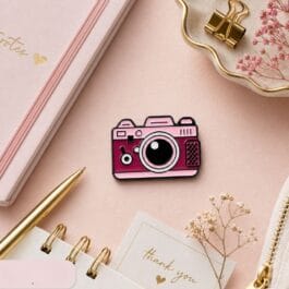 Pink camera enamel pin styled on a pastel desk with stationery and floral accents
