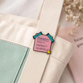 Pink reminder enamel pin with teal tape and yellow star on a light desk beside a notebook